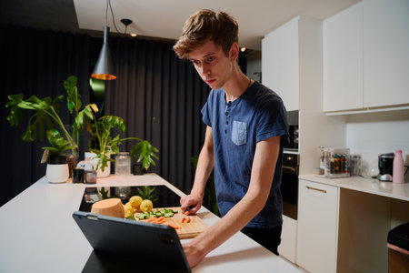 Young caucasian man in casual clothing while preparing dinner by digital tablet in kitchen at homeの写真素材
