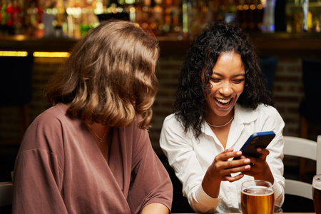 Happy young multiracial friends wearing casual clothing smiling while using mobile phone at restaurantの写真素材