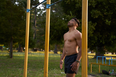 Shirtless young biracial man looking up at pull-up bar by trees in the park