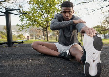 Young black man wearing sports clothing sitting and exhaling while doing stretches in parkの写真素材