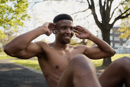 Shirtless young black man wearing wireless earphones doing sit-ups while smiling in parkの写真素材