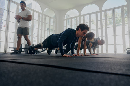 Trainer applauding group of multiracial young adults wearing sports clothing doing push-ups at the...