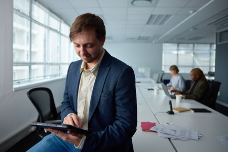 Young businessman wearing businesswear sitting on desk while using digital tablet in corporate officeの写真素材
