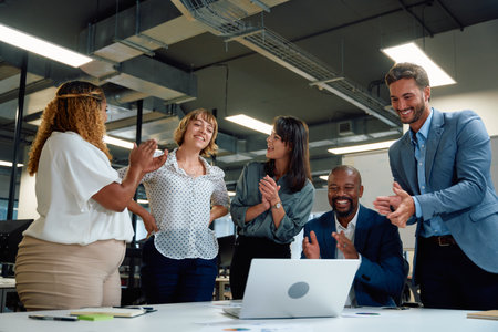 Five happy multiracial business people applauding around laptop on conference table in corporate...