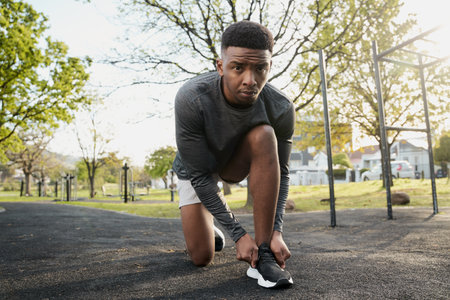Young black man wearing sports clothing looking at camera while kneeling and tying shoelace in park