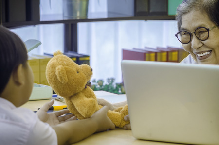 Elderly woman doctor is smiling and hands teddy bear to a sick boy. In patient's room with  laptop placed on desk.の写真素材
