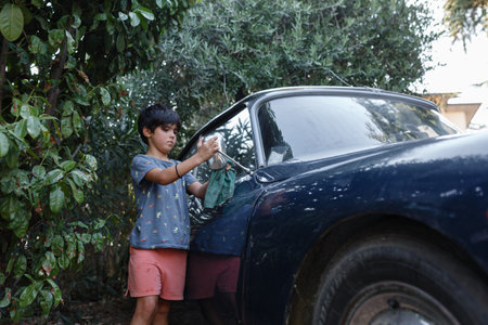 Girl of 8 years old with dark short hair washing the side mirror of blue vintage carの写真素材