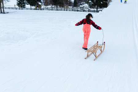 girl in red pullover and orange ski pants going uphill with wooden sled on snowy road