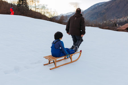 father pulling wooden sled with child with short dark hair on snowy hill against mountain backdropの写真素材