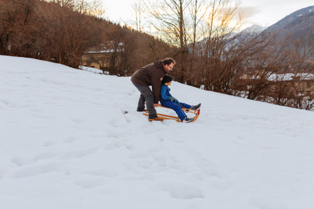 father pushing daughter sitting on wooden sled to slide down the snowy hillの写真素材