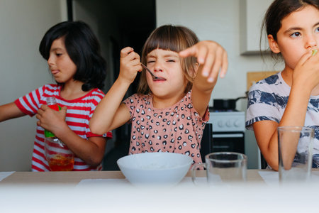 small child eating lunch at home at white table with two other children sitting on the sideの写真素材