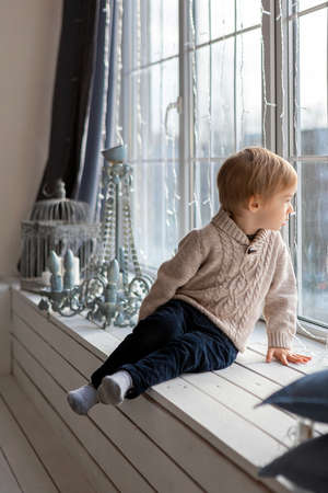 A blond boy is sitting and looking out the windowの写真素材