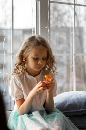 A girl cleans a tangerine sitting by the windowの写真素材