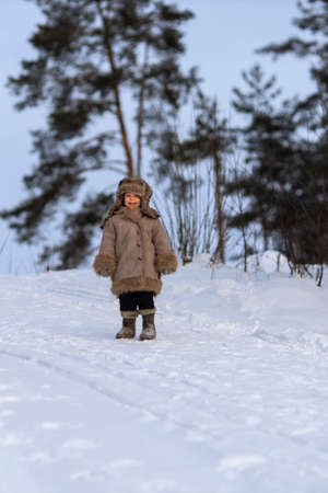 A kid in a winter fur coat, hat and felt boots in a snow-covered forestの写真素材