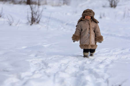 A kid in a winter fur coat, hat and felt boots in a snow-covered forestの写真素材