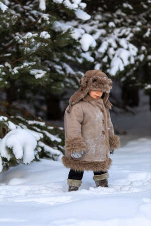 A cute boy in a fur coat and a hat with earflaps walks in winterの写真素材
