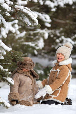 Brother and sister are sitting in the snow under a pine tree and laughing.の写真素材