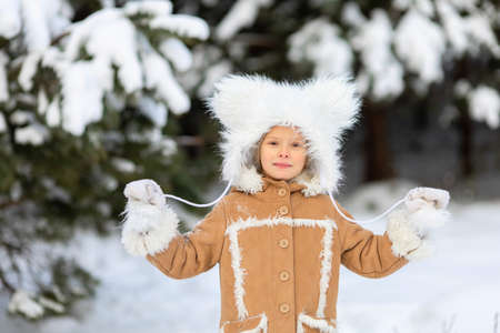 A girl in a funny fur hat with bambos in a snowy forest.The child is holding strings from a white fluffy hat in his hands.A girl in winter clothes walks and plays in a pine forest.の写真素材