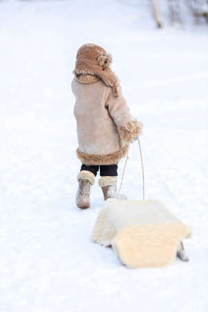 A child carries a sled by a rope on a snowy road. A boy in winter folk clothesの写真素材