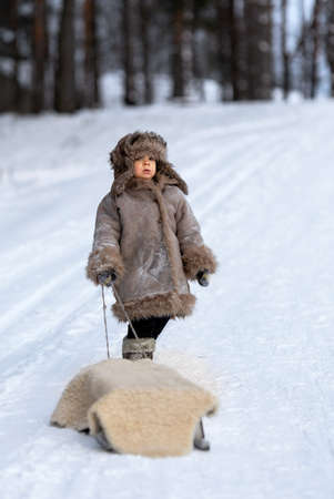 A child carries a sled by a rope on a snowy road. A boy in winter folk clothesの写真素材
