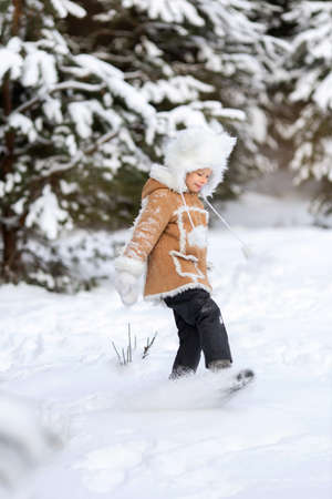 A girl in a funny fur hat and sheepskin coat is playing in a snowy forestの写真素材