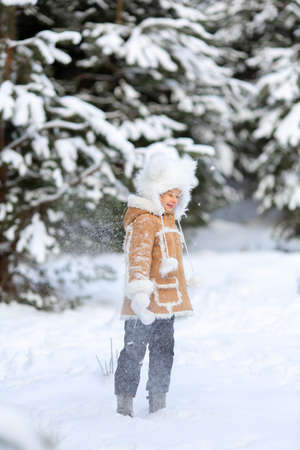 A girl in a funny fur hat and sheepskin coat is playing in a snowy forestの写真素材