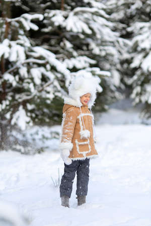 A girl in a funny fur hat and sheepskin coat is playing in a snowy forestの写真素材