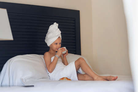 A little girl in a white towel drinks morning coffee in bed at homeの写真素材