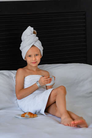 A little girl in a white towel drinks morning coffee in bed at homeの写真素材