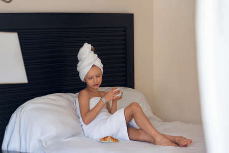 A little girl in a white towel drinks morning coffee in bed at homeの写真素材
