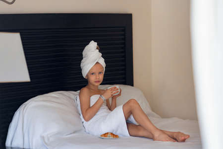 A little girl in a white towel drinks morning coffee in bed at homeの写真素材