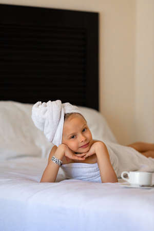 A little girl in a white towel with coffee in bed at homeの写真素材