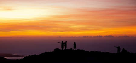 People scrutinize the horizon at sunset on top of a rock overlooking the seaの写真素材