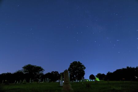 Long exposure, night photography of a the tombstones in a rural cemetery with a portion of the Milky Way faintly visible in the stars above the trees on the horizon.の写真素材