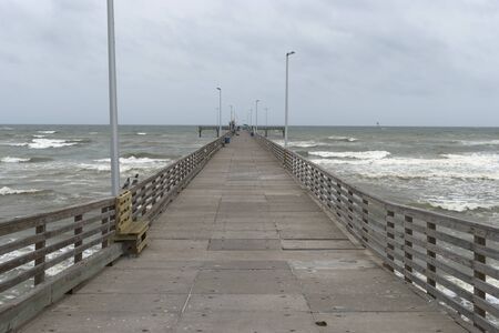 Long wood and concrete fishing pier with benches and light poles extending out into the ocean waves as they crash onto the beach.の写真素材