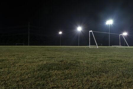 Low angle photo of a soccer goal without a net on an empty sports field at night with the lights on and the grass still wet and sparkling from a recent rain.の写真素材