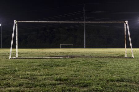 Empty soccer practice field at night looking through a goal without a net down the field to the other goal with the green grass lit up by the lights and sparkling from a recent rain.の写真素材