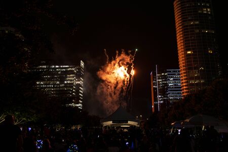 Wispy smoke clouds swirling around bright red explosions of light fill the night sky and reflect off surrounding buildings during the Independence Day Celebration at Klyde Warren Park in downtown Dallas, Texas.の写真素材