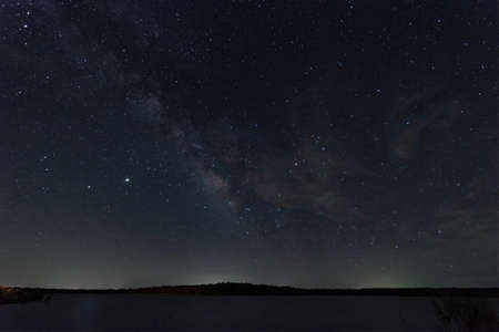 Heart of the Milky Way with some light, wispy clouds in the night sky over a lake with some trees and the glow of a town on the distant horizon.の写真素材