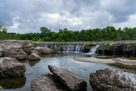 Pool filled with large rocks and boulders at the base of a rocky waterfall flowing in from a river with trees on the far shore under a cloudy, overcast sky.の写真素材