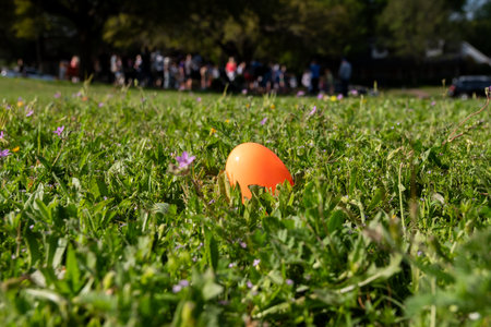 An orange, plastic Easter egg hidden in the short grass in a neighborhood park with a grup of people...