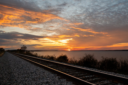 Railroad tracks leading off into the distance along the shore of a lake as the sunset fills the sky with fiery yellow and orange clouds.の写真素材