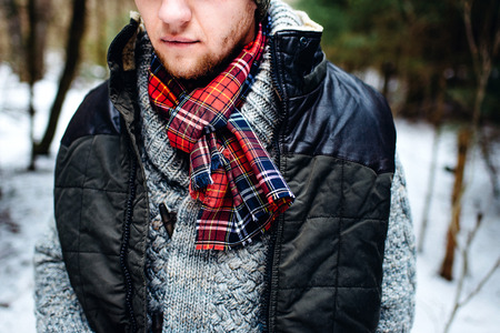 Man with a bristle standing in a cold in a warm grey sweater, black waistcoat and a red scarf with winter woods on the backの写真素材