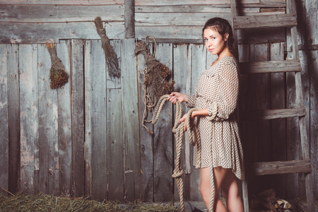 Young girl in a barn holding rope in her hands, planked wall with brooms and ladder on the back. Looking at cameraの写真素材