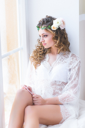 Beautiful girl in a white bridal negligee sitting next to the windowの写真素材