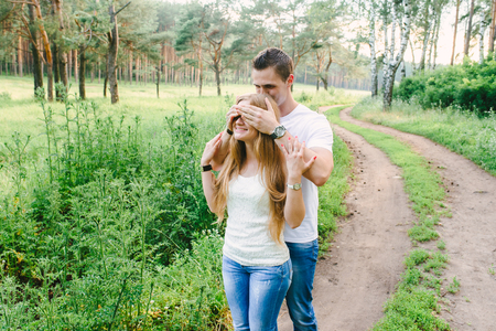 Man closing his girls eyes with his hands standing from her back, she is smiling happily and touching his hands, nature background. Man is preparing a surpriseの写真素材