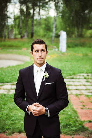 Young groom standing in a green area, hand in hand, looking worried aside, waiting brideの写真素材