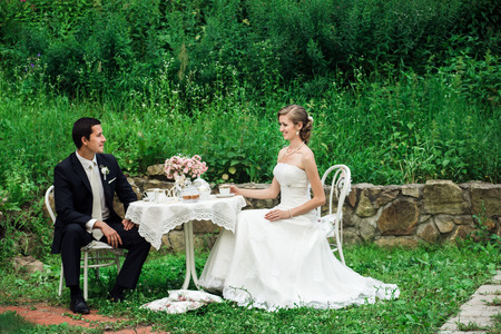 Wedding couple sitting at the table having tea, looking smiling at each other, high grass behind themの写真素材