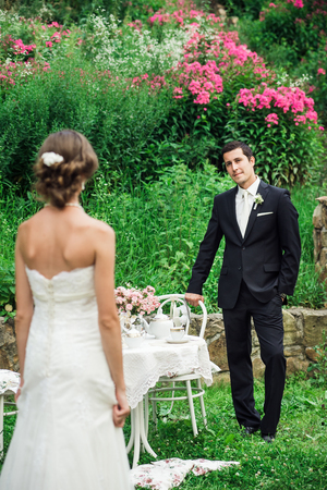 Groom in a black costume inviting his bride to sit next to him at the table, green bushes with pink flowers behind them. Picnic in the parkの写真素材
