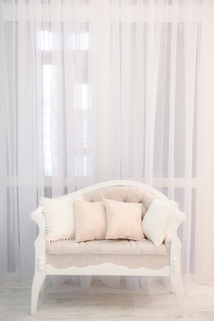 Minimalistic picture of a light beige soft sofa with nice pillow opposite light transparent curtainsの写真素材